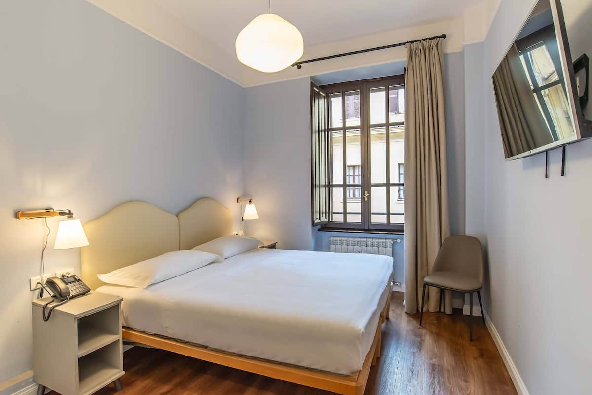 Un lit bien fait, avec une tête de lit, des lampes de chevet, un téléphone et une chaise, dans une pièce dotée d’une fenêtre et d’un radiateur.