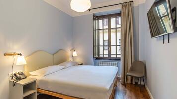 Una cama bien hecha con cabecera, lámparas de noche, teléfono y una silla en una habitación con ventana y radiador.