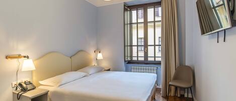 Een netjes opgemaakt bed met hoofdbord, bedlampen, een telefoon en een stoel in een kamer met raam en radiator.