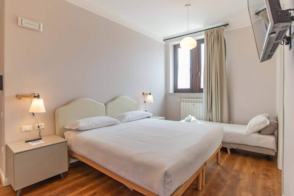 Une chambre à coucher comprenant un lit, des tables de chevet, une commode, une fenêtre avec des rideaux et un téléviseur fixé au mur.