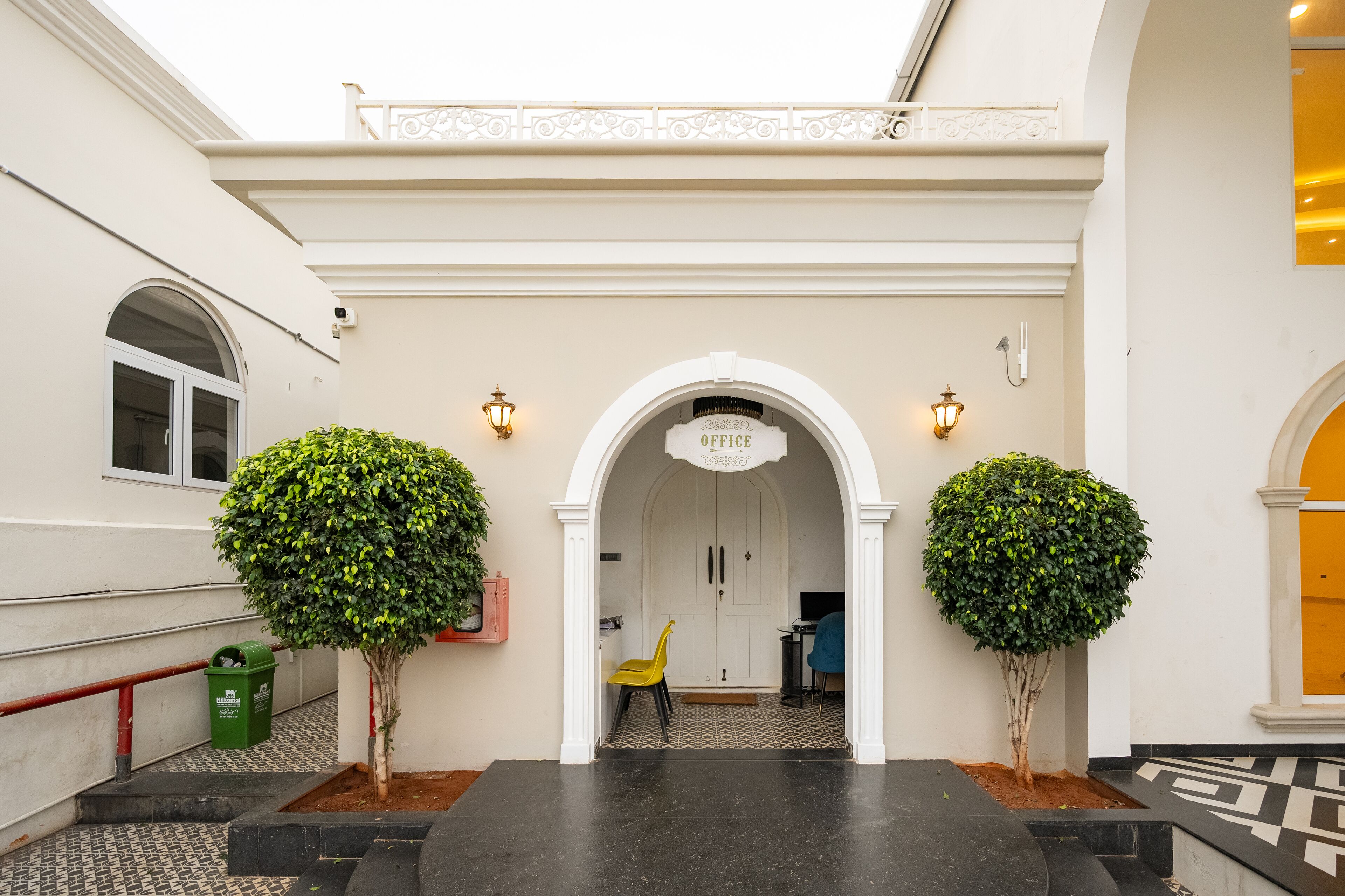 An entrance with an archway labeled "OFFICE," two potted trees, a yellow chair, and a trash bin.