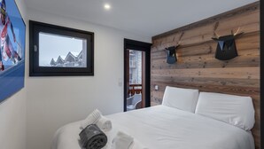Quarto com cama, almofadas brancas, janela com vista para prédios, parede de madeira com cabeças de animais montadas e uma toalha na cama.