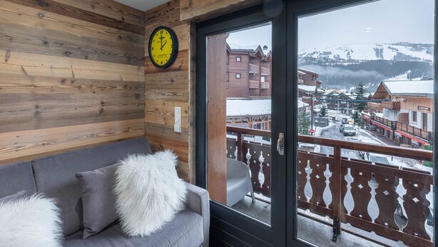 Um quarto com painéis de madeira, um sofá cinza, um travesseiro branco e fofo, e uma grande janela com vista para uma aldeia coberta de neve.