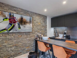 A modern kitchen with a stone wall, a large painting of a skier, and a bar area with orange chairs. - Cozy apartment for 4, near slopes in Courchevel (Courchevel 1650)