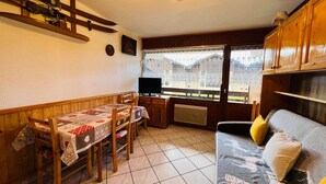 Ein gemütliches Wohnzimmer mit Essbereich, Holzmöbeln, einem Fernseher und einem Fenster mit Blick auf Gebäude.