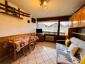 A cozy living room with a dining area, wooden furniture, a TV, and a window with a view of buildings. - Cozy studio for 4 pers with south-facing balcony (Les Saisies)
