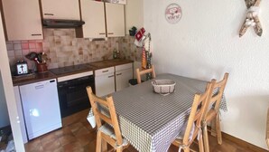 A compact kitchen with a dining area, featuring a checkered tablecloth, wooden chairs, and a small refrigerator.