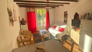 A cozy room with a bed, a dining table with chairs, a small kitchen area, and a window with red curtains.