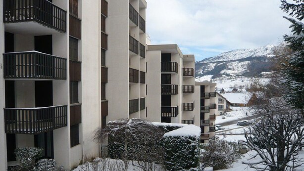 A multi-story building with balconies, surrounded by snow-covered trees and a mountainous backdrop.