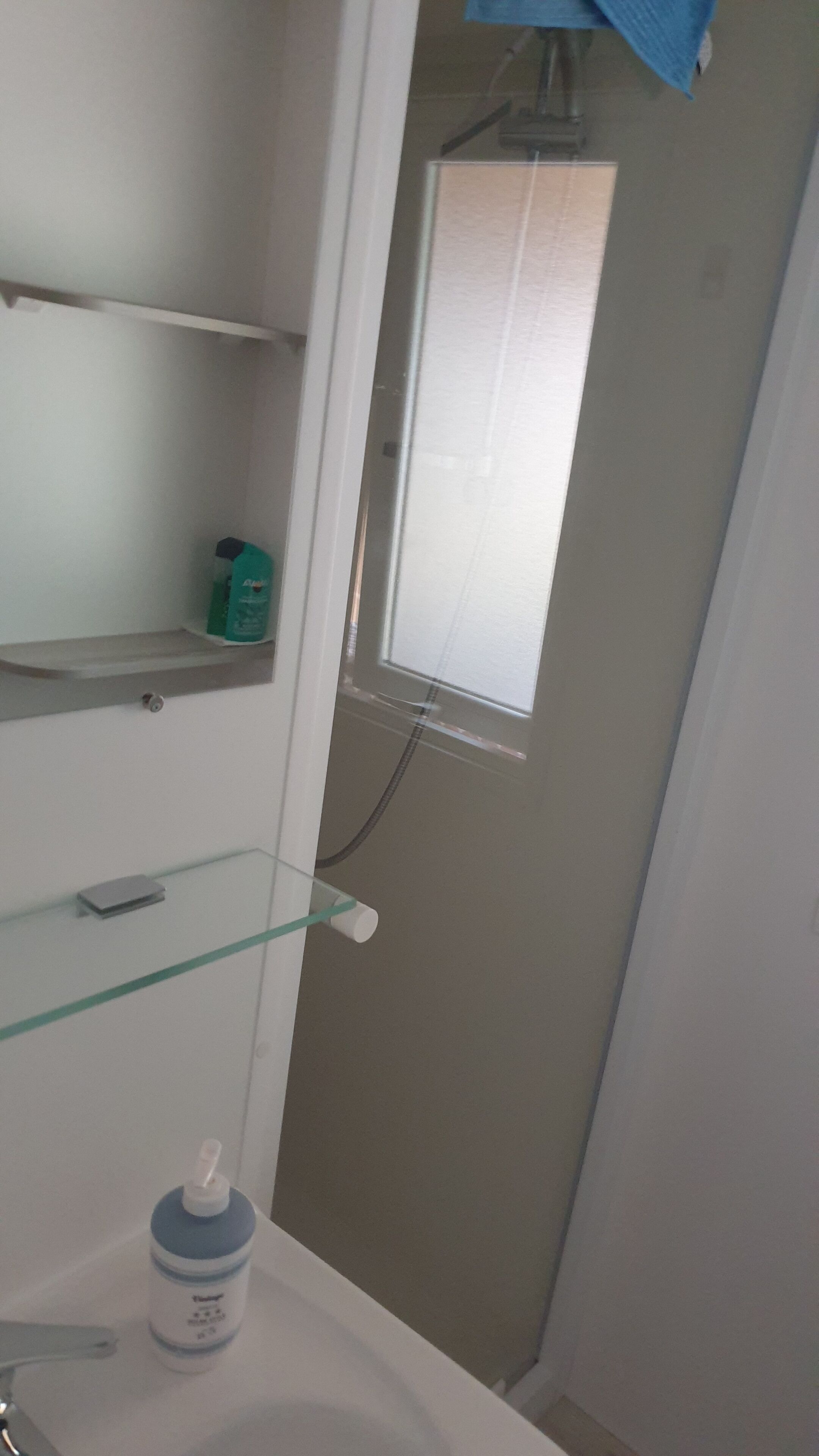 Une salle de bain avec une étagère en verre, un lavabo blanc et une fenêtre avec des stores.
