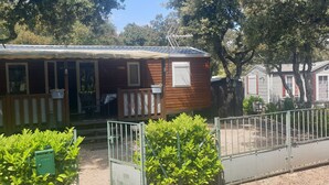 A small wooden cabin with a porch, a fenced area, and a green utility box. - Mobil-home Dans Petit Camping Tranquil. Calme, Ombragé et Nombreuses Activités (Orgnac-l'Aven)
