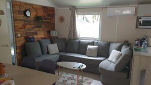 A living room with a gray sectional sofa, a small white table, and a wooden wall with a clock. - Mobil-home Dans Petit Camping Tranquil. Calme, Ombragé et Nombreuses Activités (Orgnac-l'Aven)