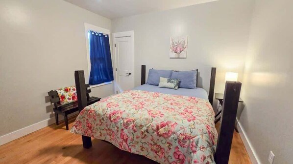 A bedroom with a bed, a nightstand, a window with curtains, a door, and a wall-mounted lamp.