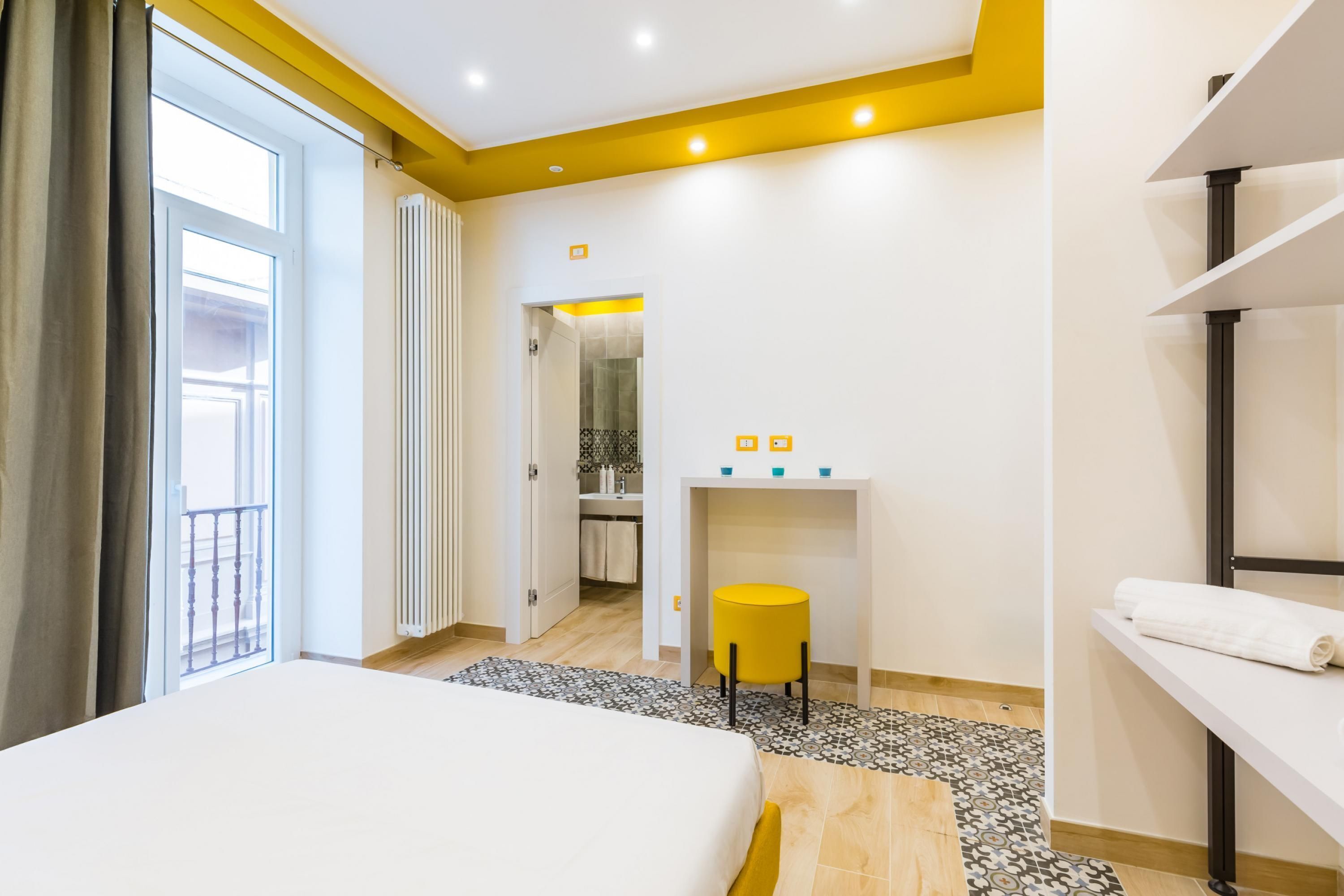 Quarto de hotel com uma cama, uma secretária pequena com uma cadeira, um banheiro visível por uma porta aberta e uma varanda com guarda-corpo.