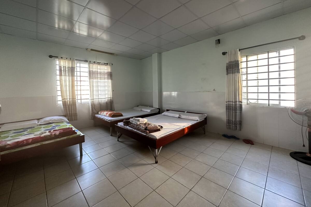 A dormitory room with three beds, tiled floors, and windows with curtains.