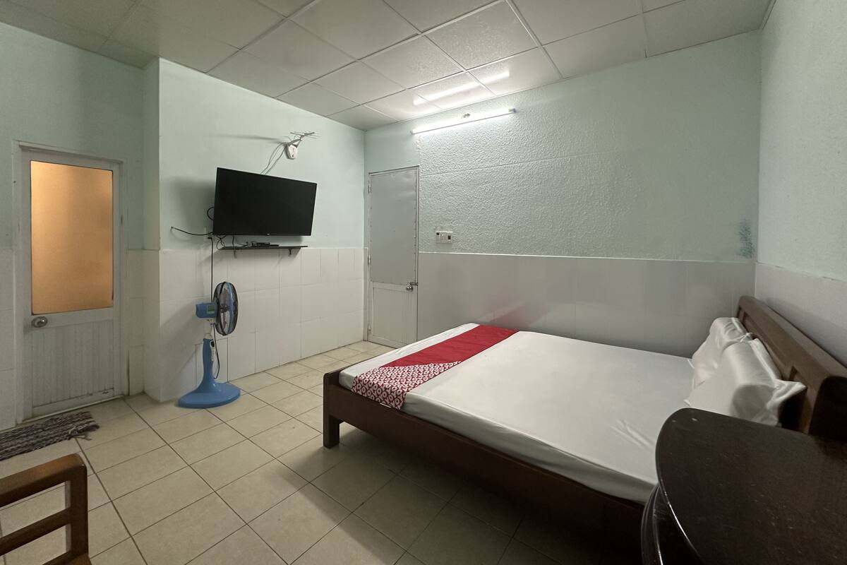 A small, simple room with a bed, a TV mounted on the wall, a fan, and a door.