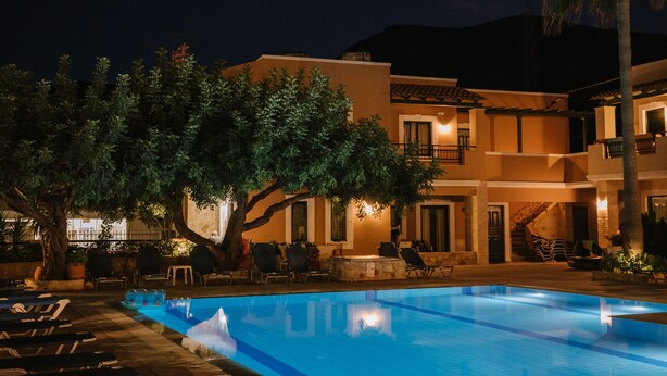 Área de piscina com espreguiçadeiras e uma árvore, cercada por um edifício com várias varandas e uma escadaria.