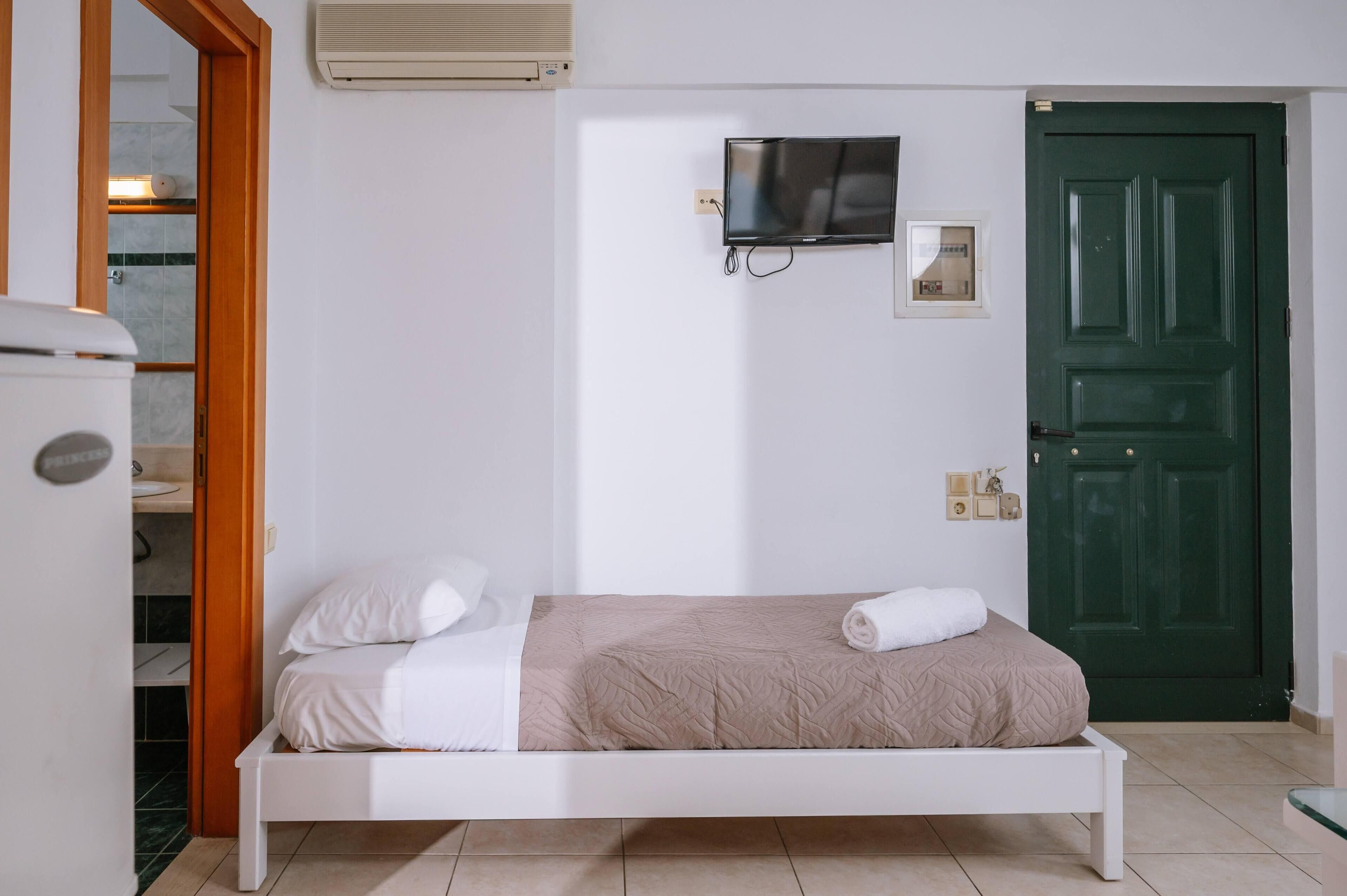 Una cama individual con una manta color beige y ropa de cama blanca, un televisor montado en la pared y una puerta verde.