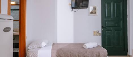 Una cama individual con una manta color beige y ropa de cama blanca, un televisor montado en la pared y una puerta verde.
