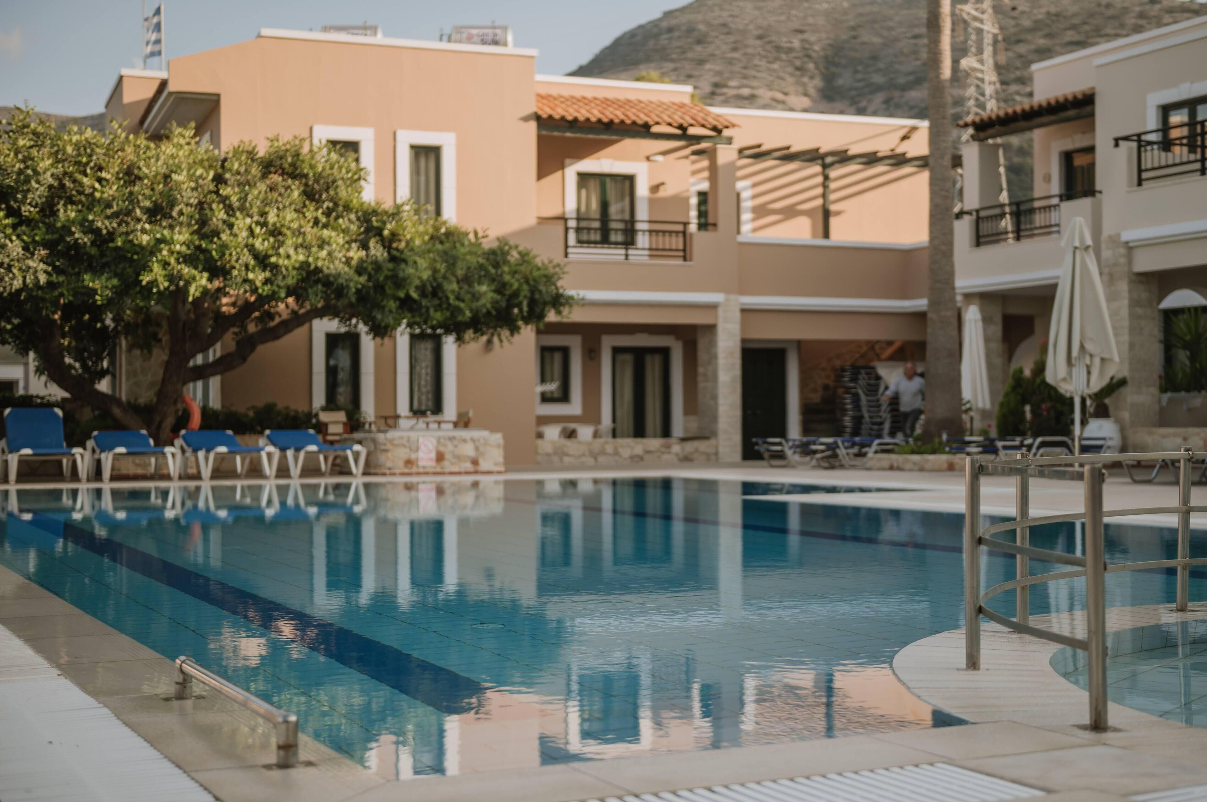 Una piscina con sillas de descanso y sombrillas, rodeada de edificios con balcones y un árbol.