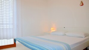A simple, clean bedroom with a bed, white walls, a nightstand with a lamp, and a window with sheer curtains. - For Your Getaway Near the Beach-beahost Rentals (San Michele al Tagliamento)