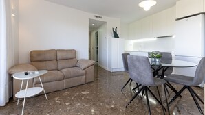 Una sala moderna con un sofá color beige, una mesita blanca pequeña y un comedor con dos sillas grises y una mesa de vidrio.