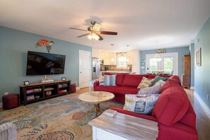 A living room with a red sofa, a television, a ceiling fan, and a kitchen in the background. - The Gypsy Rose: Private Pool - 5 Minute Walk from Beach and Splash Pad! (Oak Island)