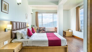 A hotel room with a large bed, a wooden nightstand, a chair, and a view of the mountains through the window. - Hotel Blue Poppy (Gangtok)