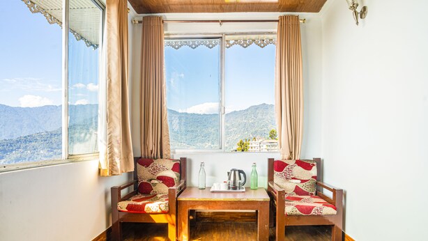 A room with two wooden chairs, a small table, and a view of mountains through large windows. - Hotel Blue Poppy (Gangtok)