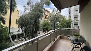 A balcony with a table and chairs, overlooking a street with buildings and trees. - Koukaki Cozy Studio Retreat 1' from metro (Athina)