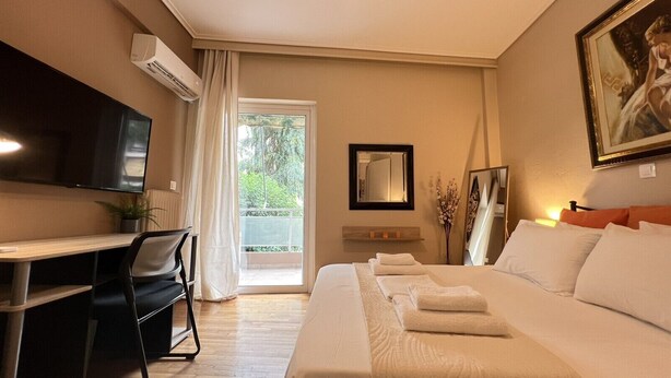 A hotel room with a bed, a desk, a chair, a TV, a balcony with a view of greenery, and a framed picture on the wall. - Koukaki Cozy Studio Retreat 1' from metro (Athina)