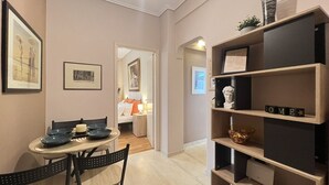 A modern interior with a dining area featuring a round table and chairs, a bookshelf with decorative items, and a cozy bedroom visible through an open door. - Koukaki Cozy Studio Retreat 1' from metro (Athina)