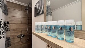A bathroom with a wooden shower enclosure, a mirror, and a shelf with multiple bottles of Karavaki shower gel. - Koukaki Cozy Studio Retreat 1' from metro (Athina)