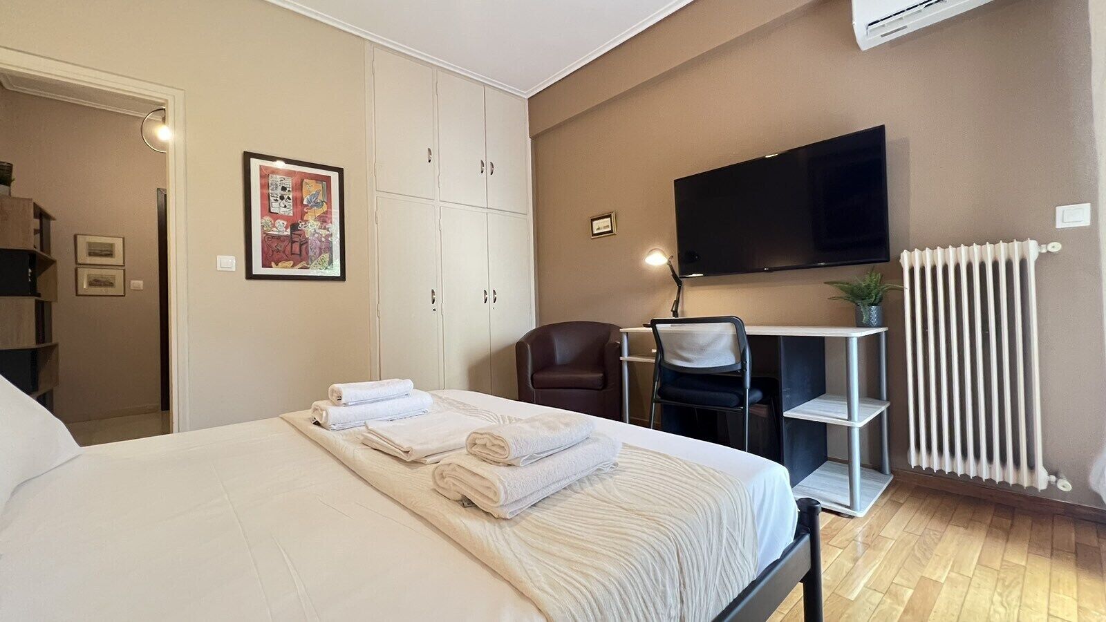A hotel room with a bed, a desk, a chair, a TV, and a radiator.