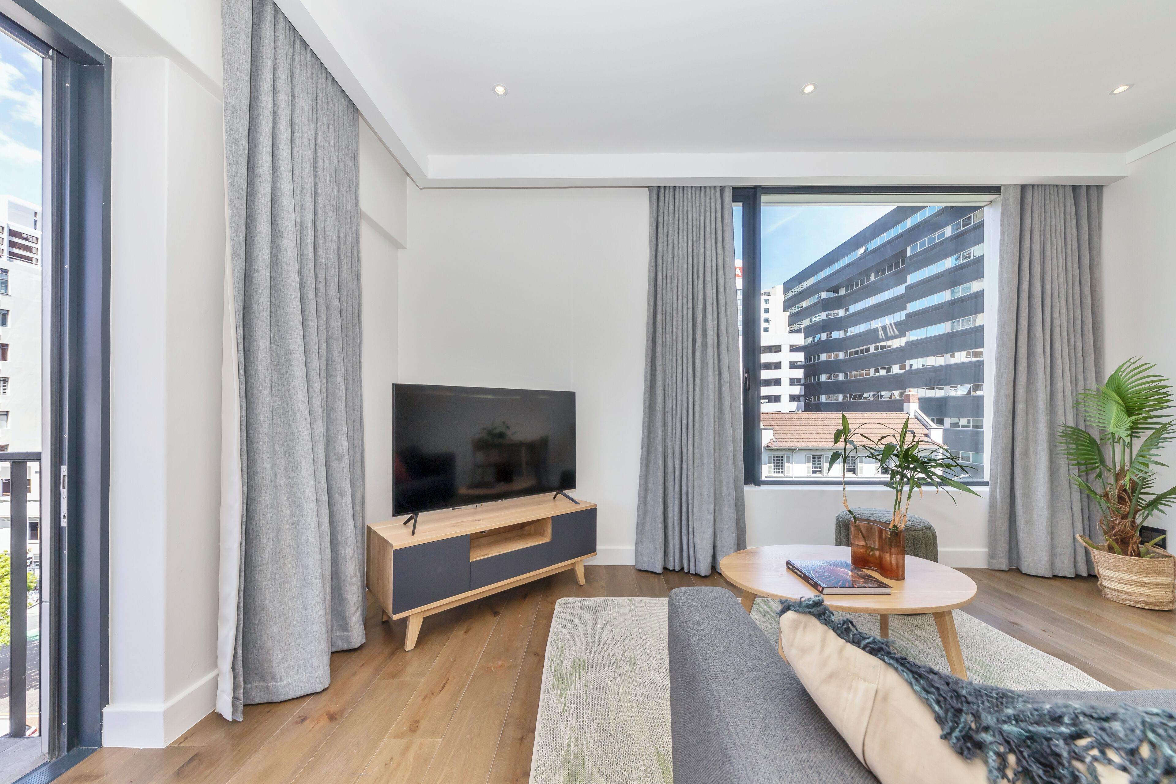A modern living room with a flat-screen TV, grey curtains, a sofa, a coffee table, and a view of a building through large windows.