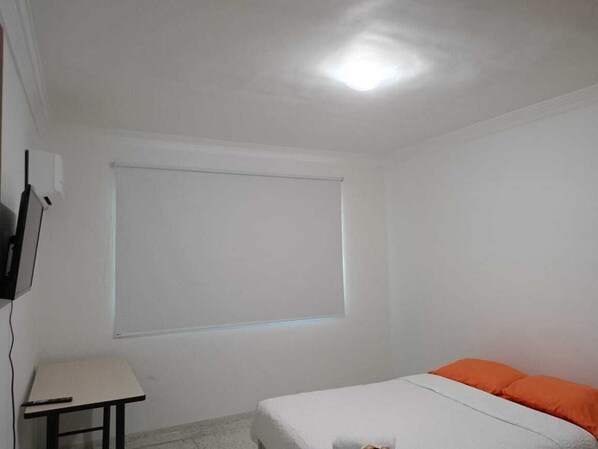 A room with a bed, a small table, a TV mounted on the wall, and a window with blinds.