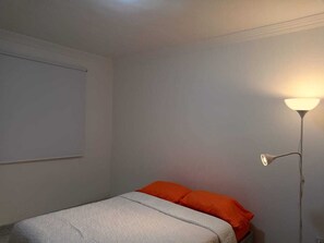 A single bed with an orange pillow, a white blanket, and a floor lamp with a white shade.