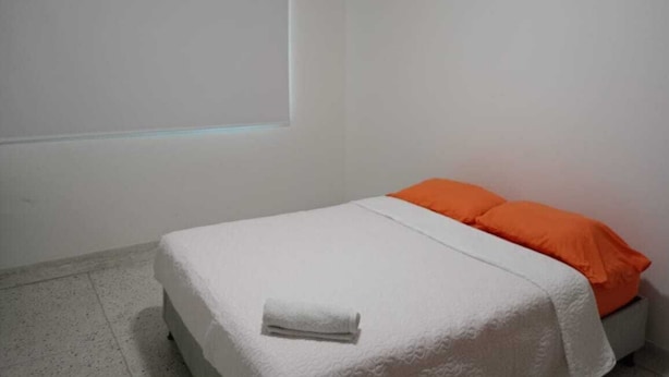 A single bed with a white bedspread and an orange pillow, a white towel on the bed, and a window with blinds.