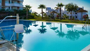 Piscina com escada de metal, palmeiras e um edifício com varandas ao fundo.