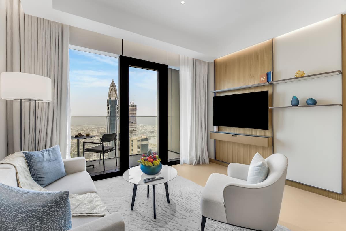 A modern hotel room with a large window offering a city view, a flat-screen TV mounted on the wall, and a comfortable seating area with a coffee table.