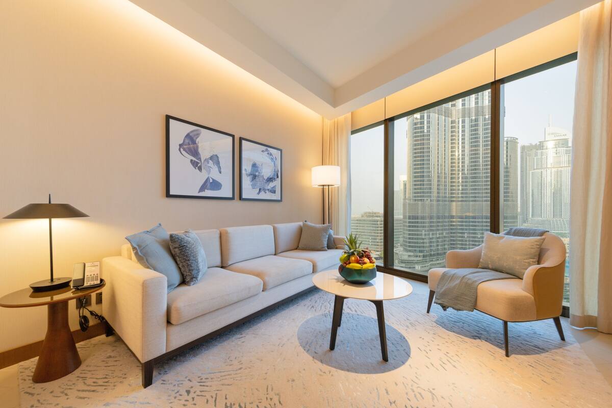 A modern hotel room with a sofa, armchair, and coffee table. There are two framed artworks on the wall, a lamp, and a view of the cityscape through large windows.