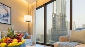 A modern hotel room with a large window offering a city skyline view, a round table with a fruit bowl, a sofa, and a lamp. - EDEN'S Homes & Villas - Address Opera District Residences (Dubai)