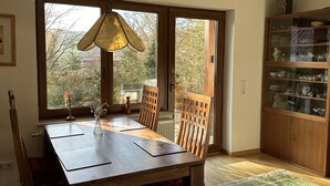 Een eetruimte met een houten tafel en stoelen, een hangende lamp en een uitzicht door een glazen deur naar buiten.