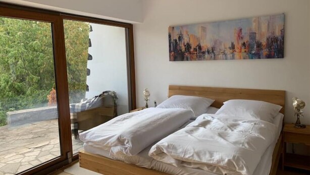 Een slaapkamer met een houten bed, wit beddengoed, een stadsgezichtschilderij aan de muur en een schuifdeur naar een buitenruimte.