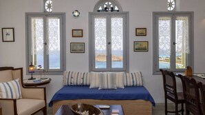 Una habitación acogedora con una cama, una mesita con un cuenco, y una silla. Hay dos ventanas con cortinas, una lámpara de pared y cuadros enmarcados.
