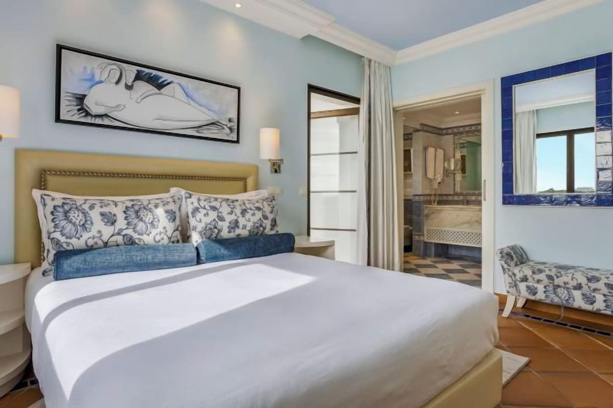 A hotel room with a large bed, a headboard, a nightstand, a wall-mounted lamp, a painting, a bathroom visible through an open door, and a window with blue tiles.
