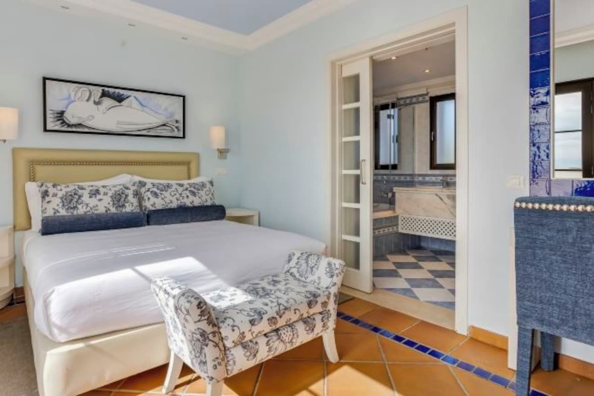 A bedroom with a bed, a chair, a painting, and a bathroom visible through an open door.