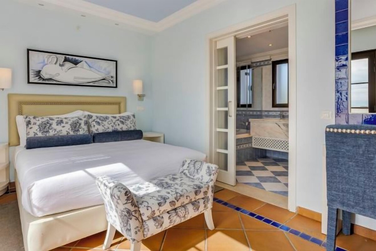 A bedroom with a bed, a chair, and a bathroom visible through an open door.