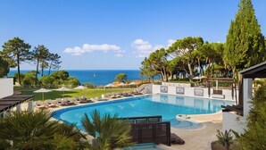Zona de piscina amb gandules i ombrelles, envoltada d'arbres i vista al mar.
