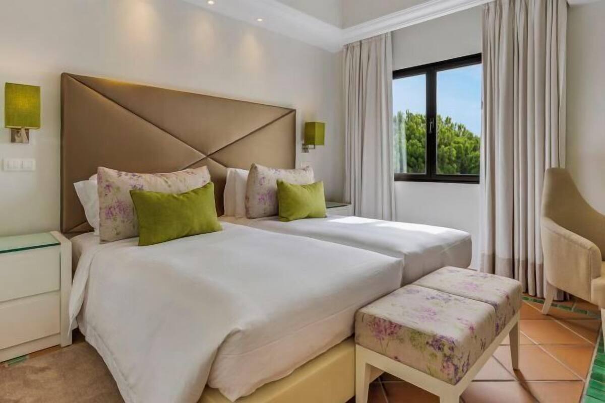 A hotel room with two beds, a bench, a chair, and a window with curtains.